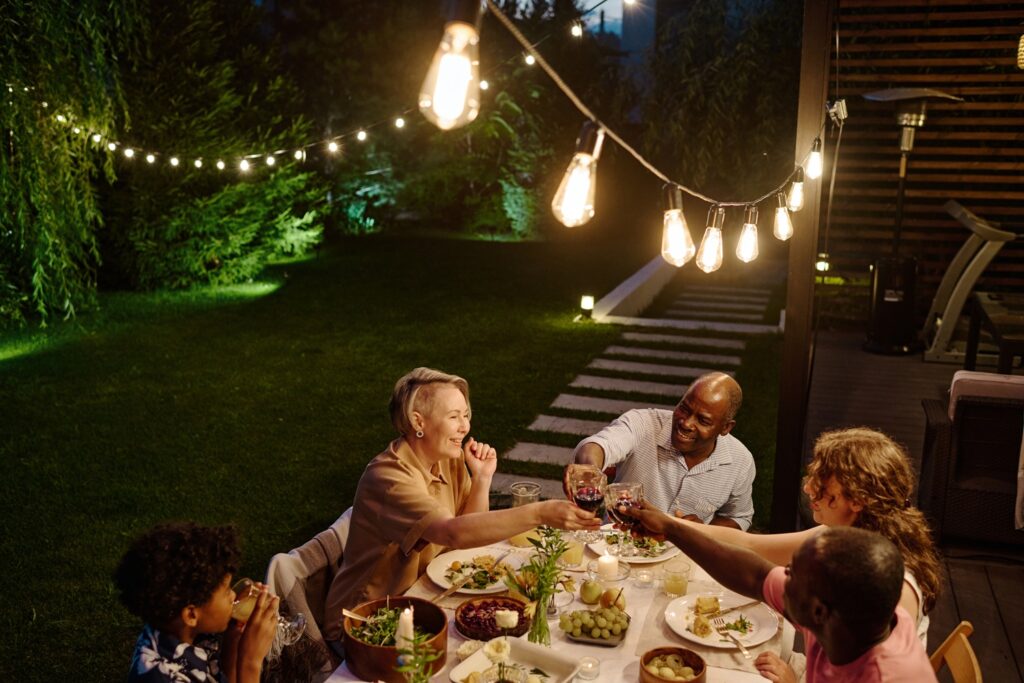 string lights illuminating large intercultural family toasting with glasses of wine over table served with variety of food while having dinner