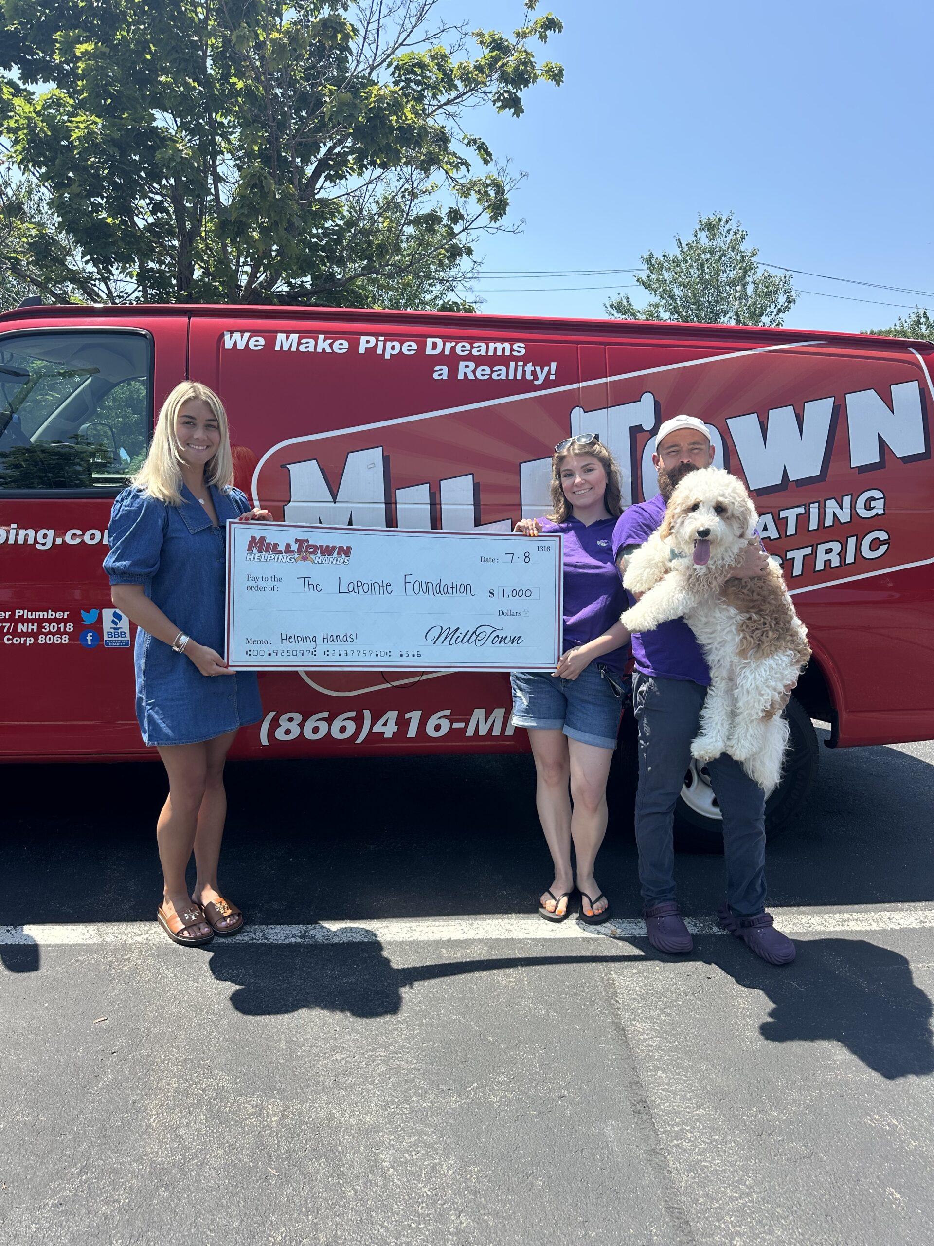 Three people and a fluffy dog pose with a giant check in front of a red MilTown promotional van.