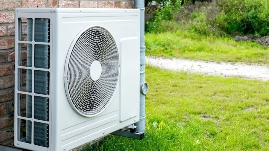 Outdoor air conditioner condenser unit mounted on a brick wall beside a grassy yard. The large fan is visible through a protective grille.